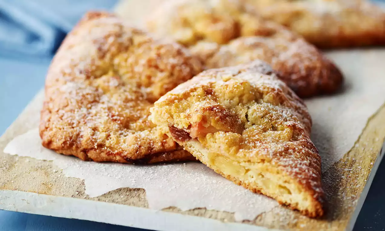 Gyldne trekantede æbletasker med let sprød krumme, drysset med flormelis og lagt på et lysebrunt skærebræt