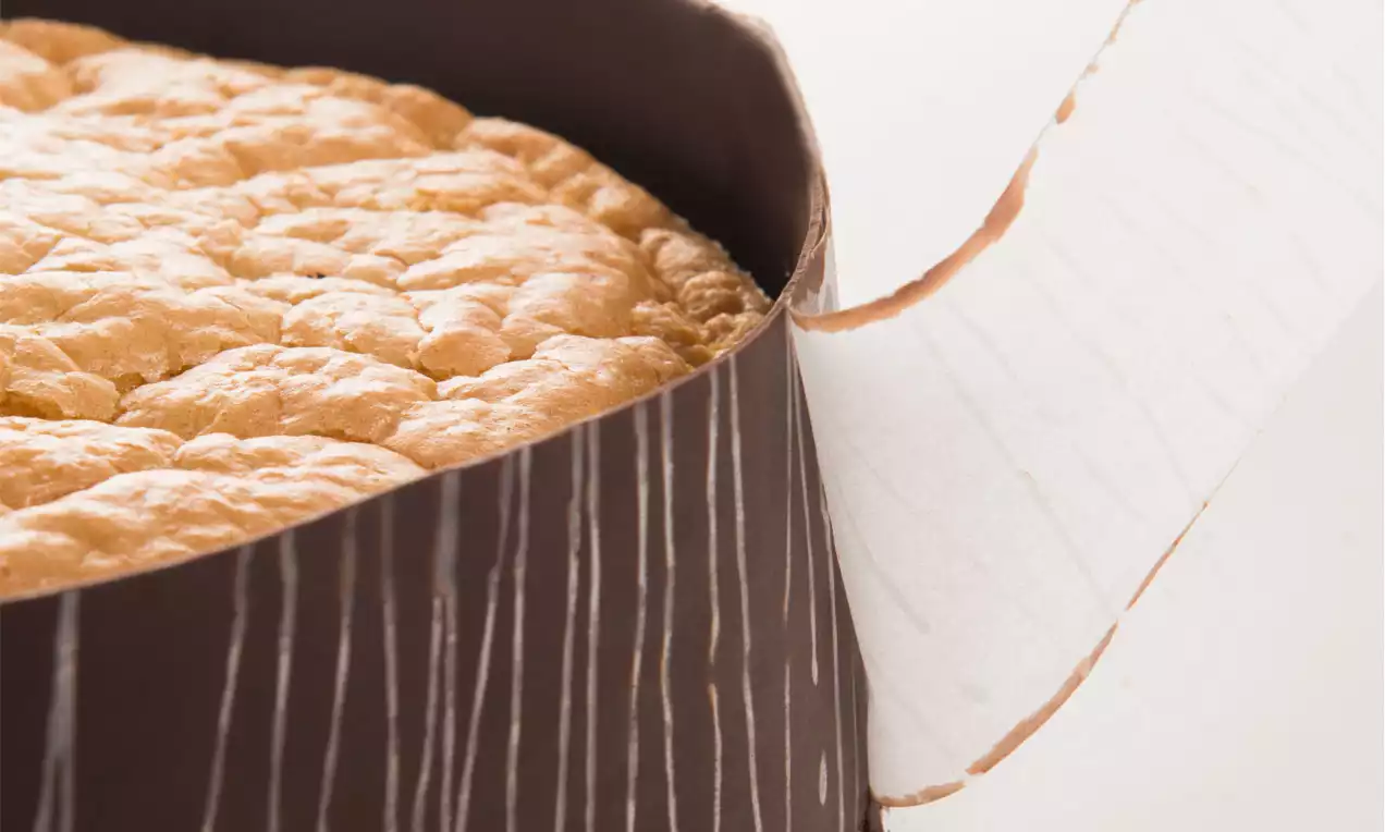 Goldbrauner Biskuit mit luftiger Struktur, umhüllt von dunkelbraunem Schokoband mit weißen Streifen und leicht gelöstem Pergamentpapier