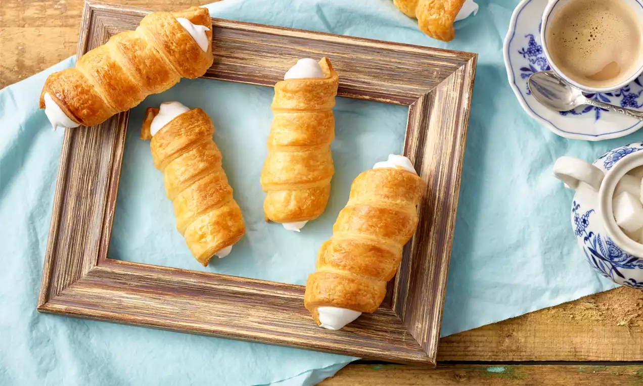 Schaumrollen aus goldbraunem, gebackenem Topfenteig mit glänzender Oberfläche, gefüllt mit weißer Creme, serviert in einem Holzrahmen