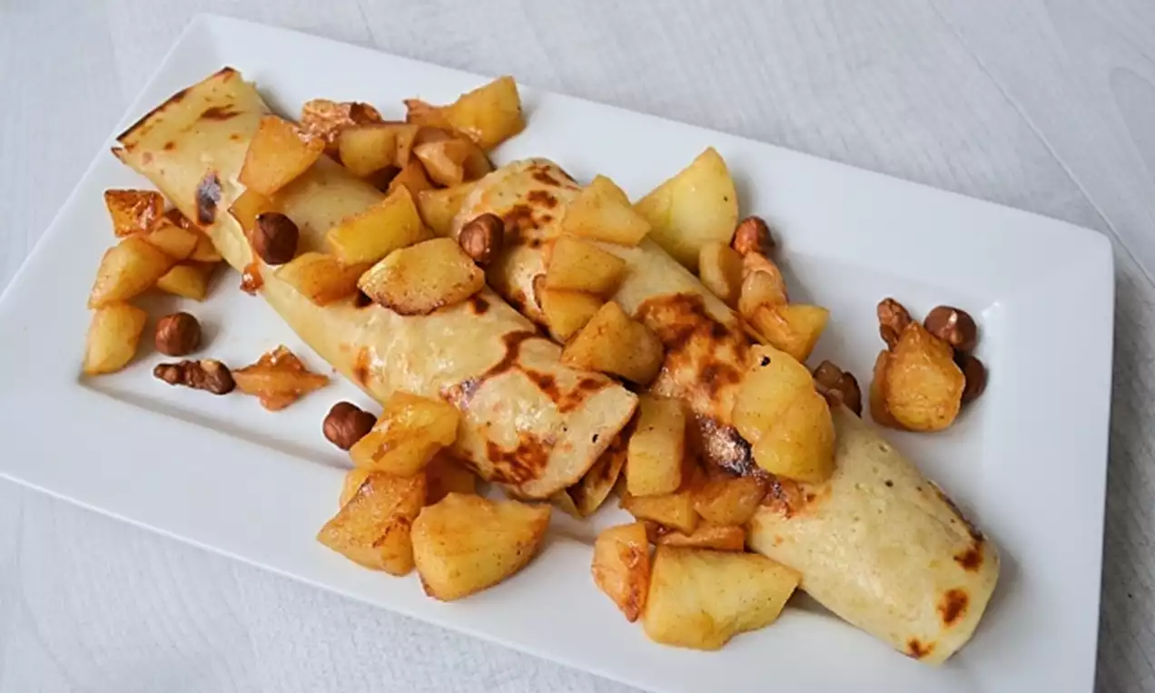 Crêpes roulées dorées garnies de morceaux de pommes caramélisées et noisettes dispersées sur une assiette blanche
