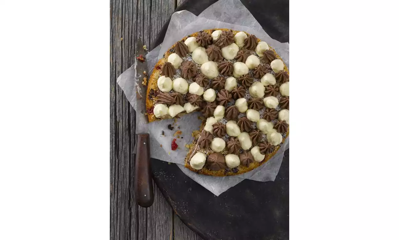 Pyöreä kakku leivinpaperilla, täynnä vaaleita ja tummia mousse-pursotuksia, reunoilta hieman mureneva, leikattu viipaleella näkyville