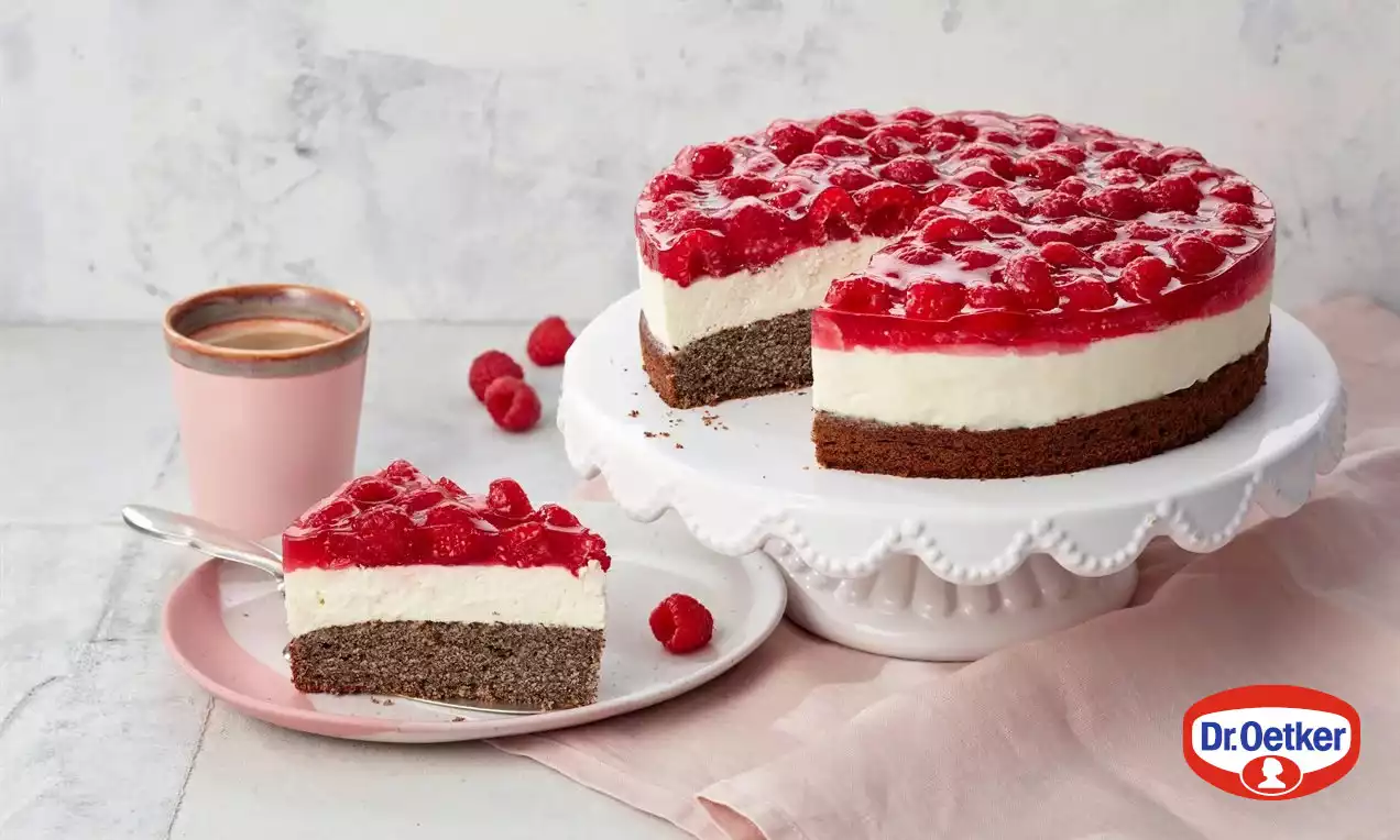 Makový tort s tmavohnedým základom, strednou vrstvou bieleho krému a lesklým červeným malinovým vrchom na bielom stojane, vedľa šálky kávy na ružovej servítke