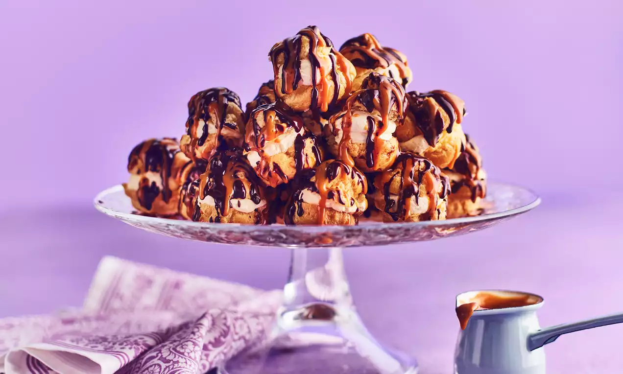 A pyramid of golden choux bites drizzled with glossy chocolate and caramel, displayed on a clear glass stand with a lilac backdrop