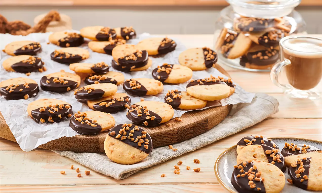 Runde, goldbraune Kekse mit glänzend dunkler Schokoladenglasur und knusprigem Krokant auf Holzbrett mit Backpapier und Glas voller Kekse