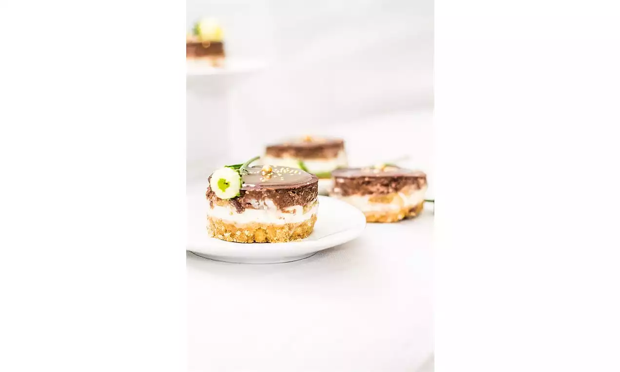 Mini cheesecake s křupavou sušenkovou základnou, krémovou vrstvou a čokoládovým vrškem, zdobený kvítkem na bílém talíři proti světlému pozadí