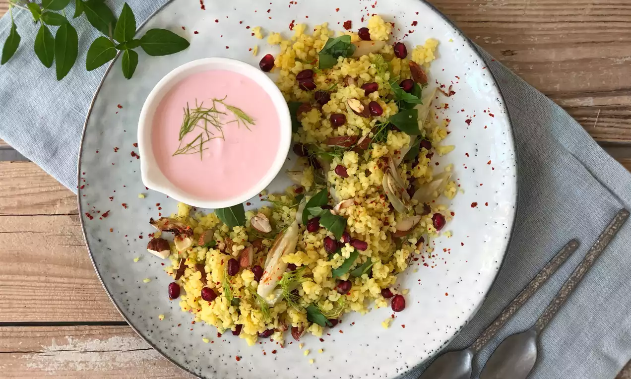Bulgursalat in leuchtendem Gelb mit roten Granatapfelkernen, Kräutern und Mandelsplittern auf hellem Teller neben rosa Joghurtdip