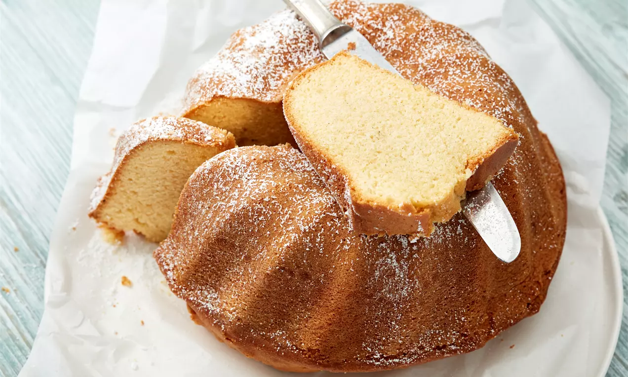 Lockerer Vanille-Gugelhupf mit goldener Kruste und Puderzucker bestäubt, ein Stück herausgeschnitten auf weißem Teller mit Vanilleschote