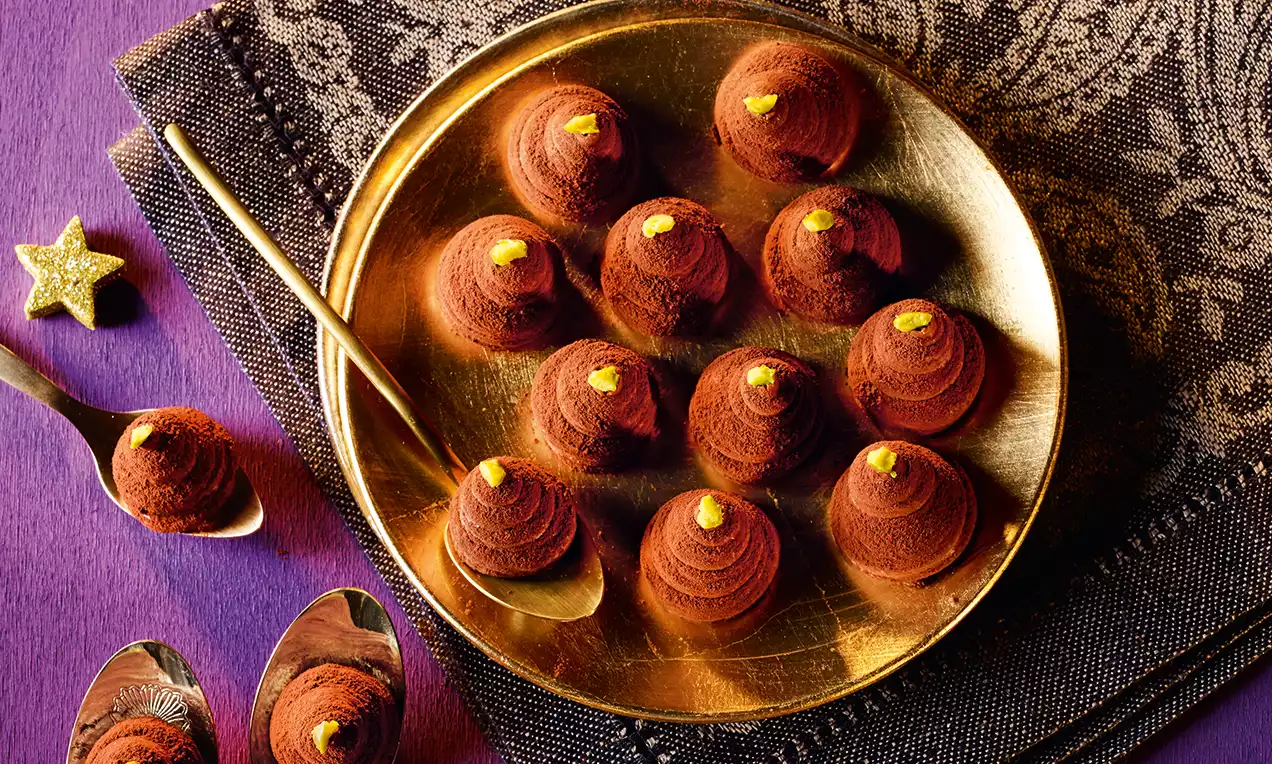 Altın rengi tabakta kakao serpilmiş, spiral şekilli çikolatalı tatlılar, üzerlerinde sarı süslemeler ve yanında metal kaşıklar