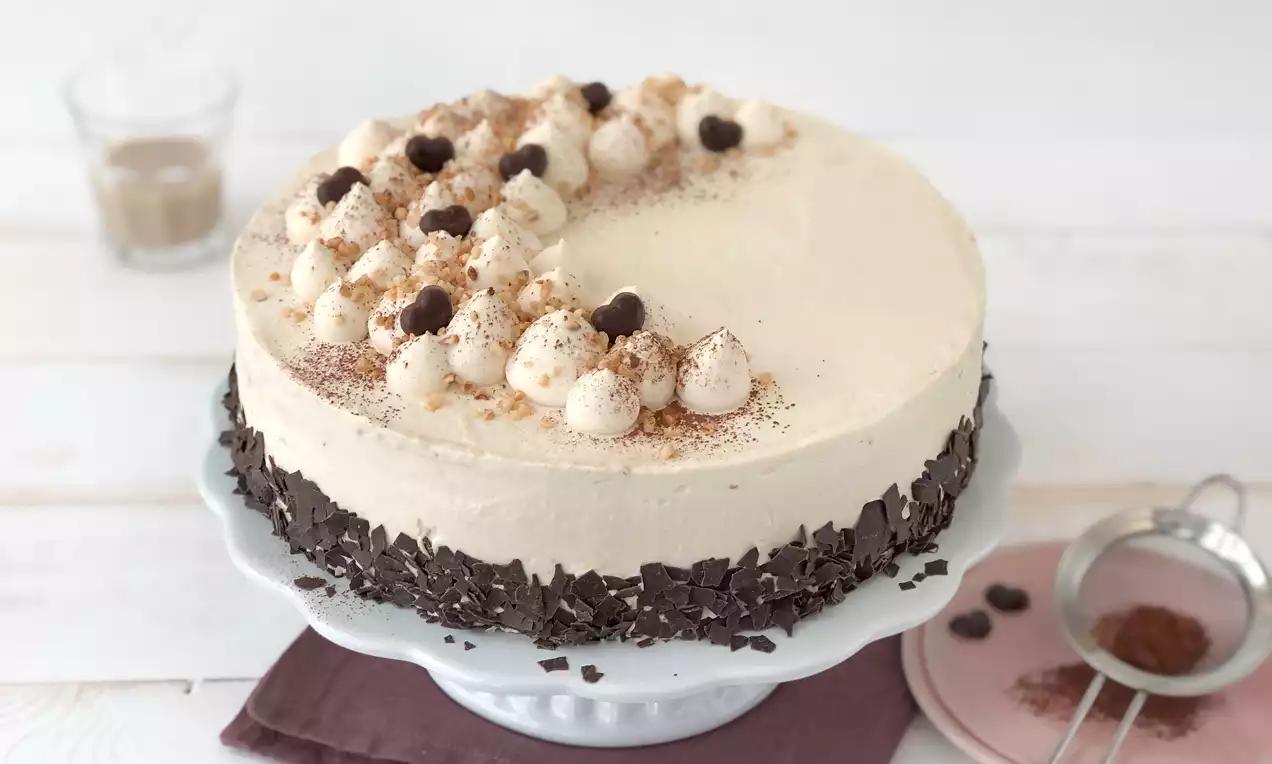 Baileys®-Torte mit zarter Creme und Schokoraspeln am Rand, oben Sahnetupfen, Kakaopulver, Schokodrops auf weißer Tortenplatte