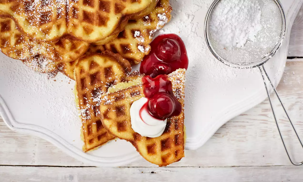 Goldbraune, herzförmige Waffeln mit zarter Puderzuckerschicht, dekoriert mit roten Kirschen und Sahne auf weißer Servierplatte