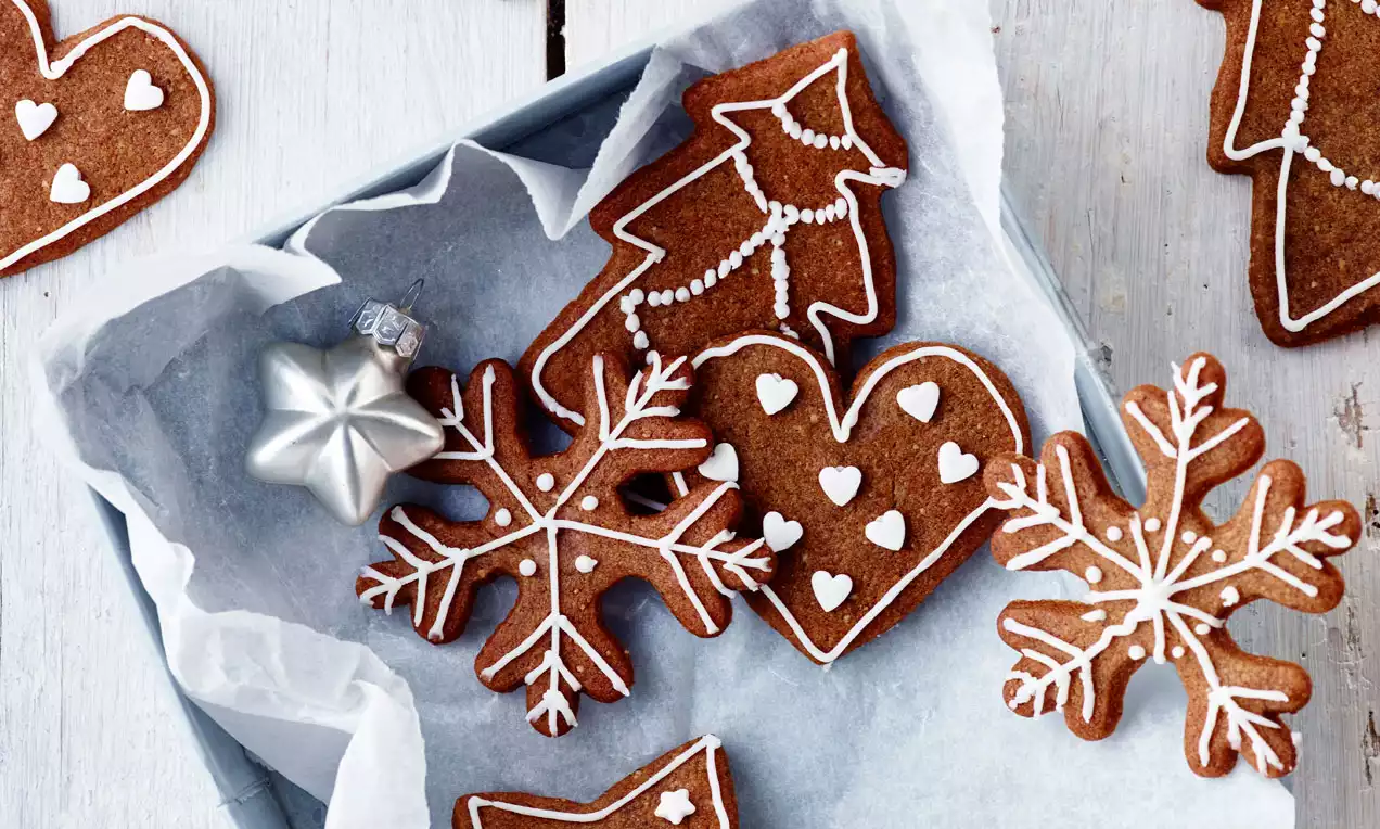 Brunkager formet som stjerner, hjerter og grantræer dekoreret med hvid glasur og placeret i en bakkefor med pergamentpapir 