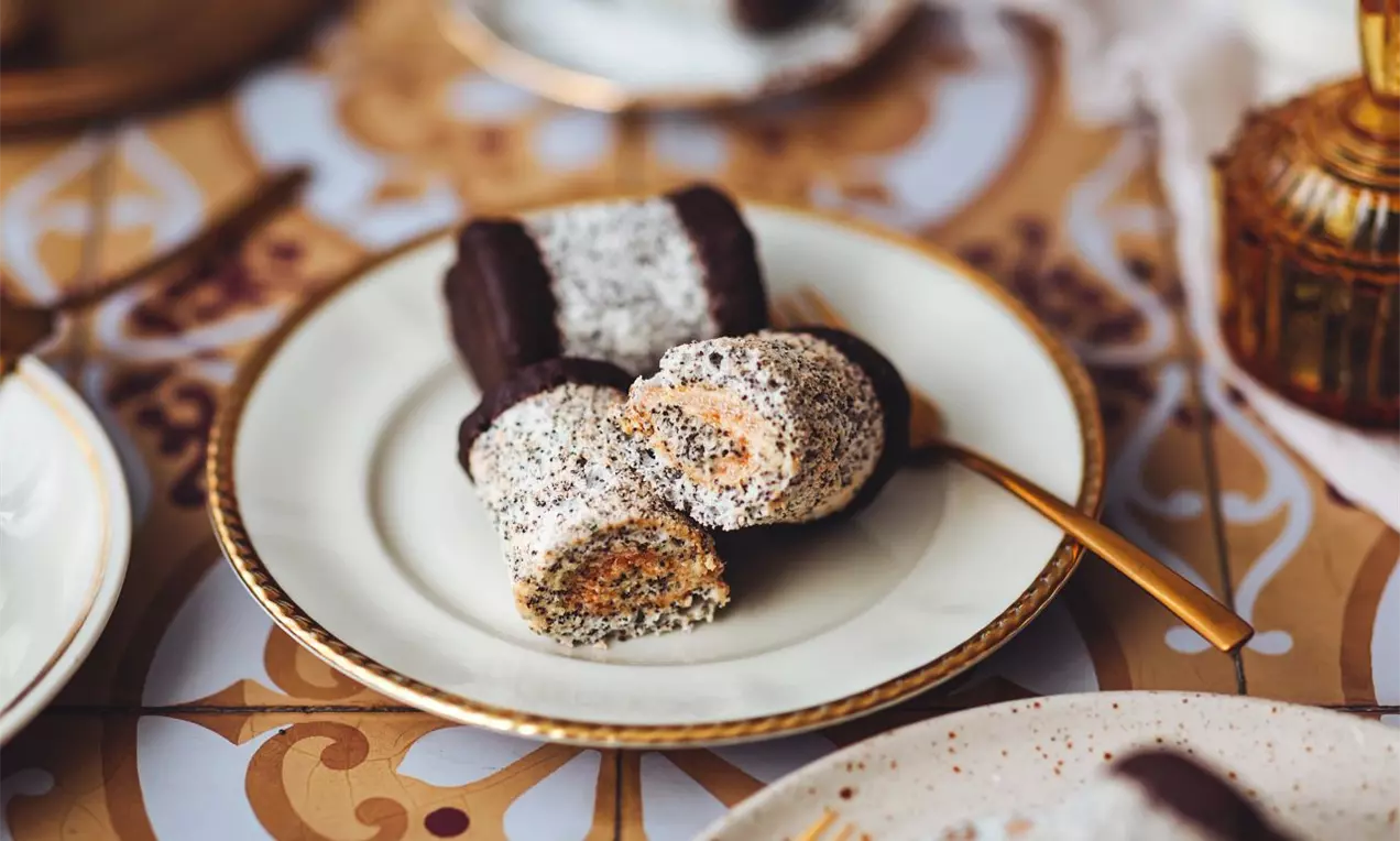 Makový rolovaný múčnik plnený hnedým karamelovým krémom, poliaty tmavou čokoládou na koncoch na tanieriku so zlatým okrajom a zlatou vidličkou