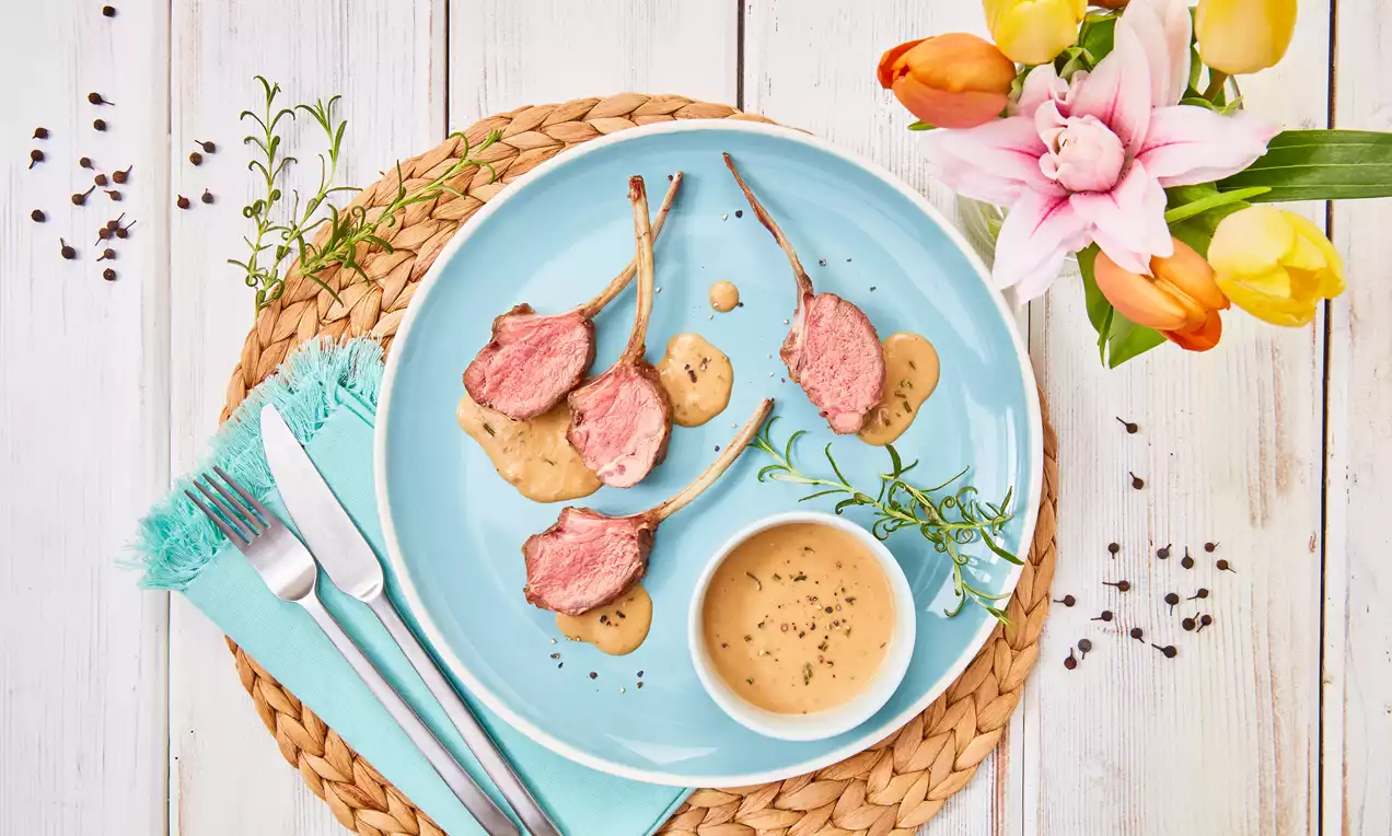 Zartrosa Lammkarrees mit goldbrauner Kruste auf heller Sauce, angerichtet auf einem blauen Teller neben Kräutern und bunter Blumendekoration
