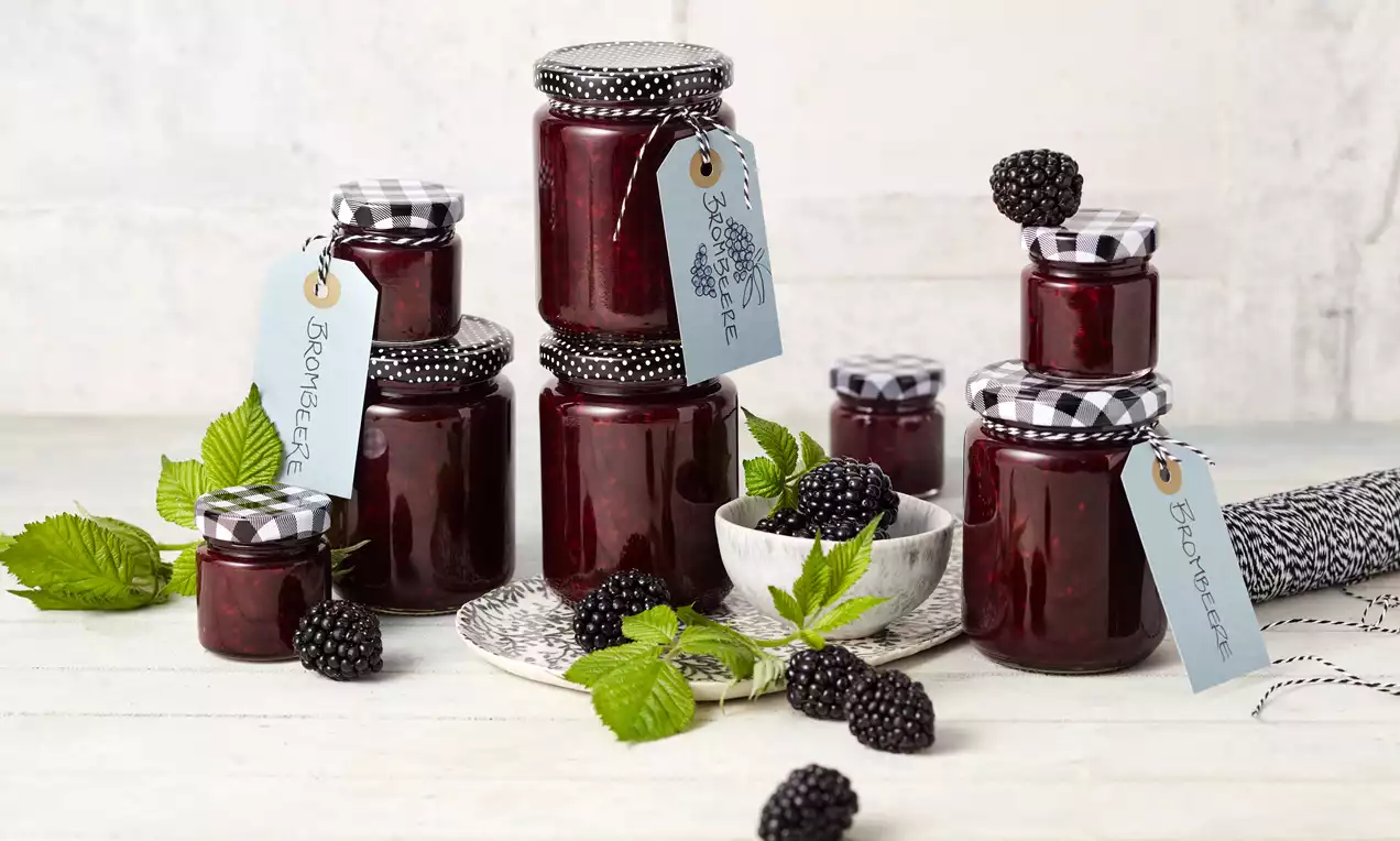 Gläser mit dunkelvioletter Brombeermarmelade, teils mit kariertem Stoffdeckel und Etikett, umgeben von frischen Brombeeren und grünen Blättern