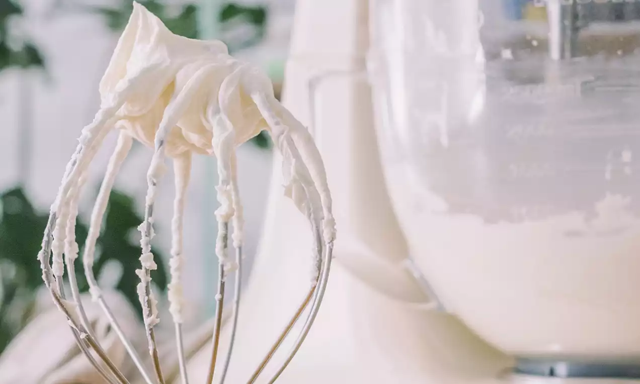 Crema de mantequilla blanca y suave con textura aterciopelada adherida a un batidor metálico frente a una batidora blanca