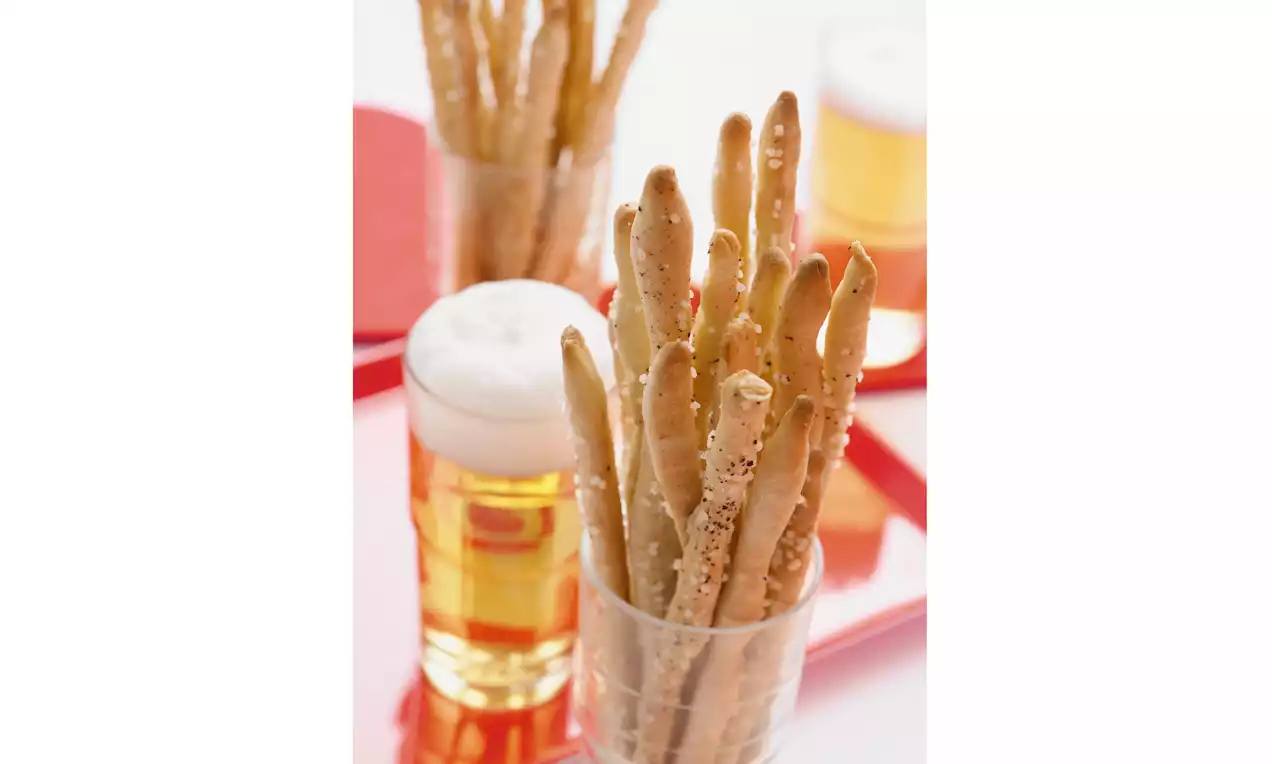 Grissini dorati e sottili con una superficie croccante e punteggiata di semi, disposti in un bicchiere accanto a un boccale di birra effervescente