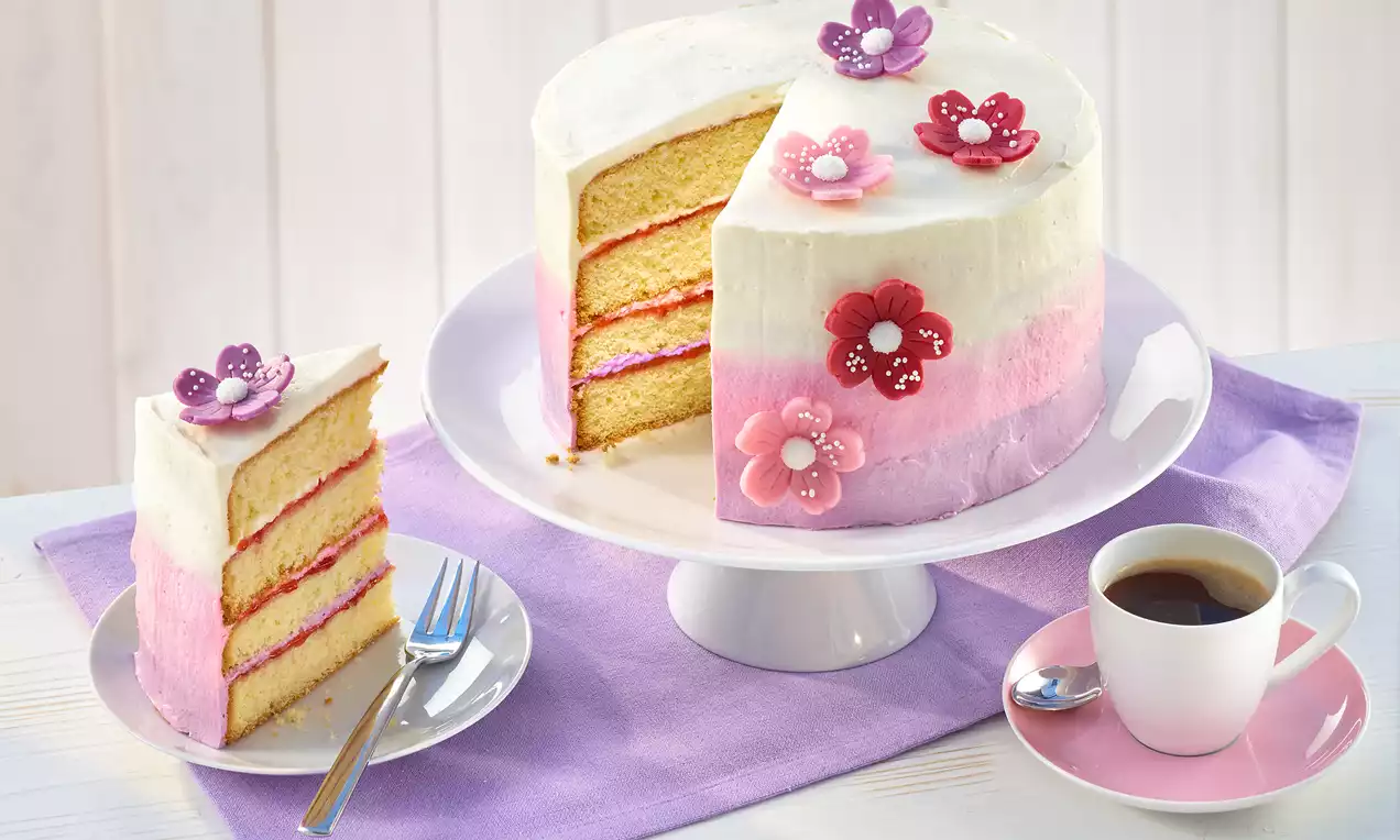 Blumen-Torte mit Ombré-Glasur in Weiß, Rosa und Pink, verziert mit Blüten aus Zucker, auf weißer Tortenplatte mit violettem Tuch drapiert