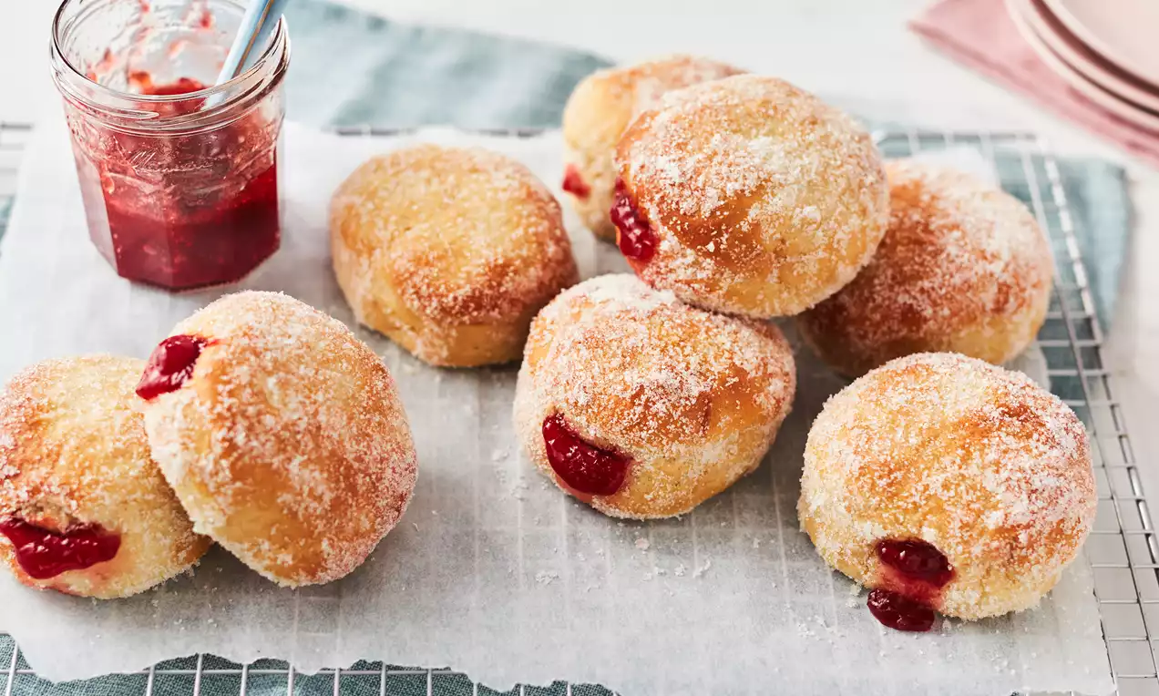 Golden sugar-dusted doughnuts with soft round shapes, oozing red jam filling, arranged on parchment over a cooling rack