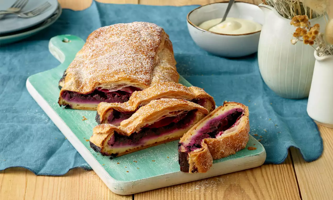 Goldbrauner Strudel mit fluffigem Teig und dunkler Schwarzbeerfüllung, angeschnitten auf hellblauem Holzbrett mit Puderzucker bestäubt