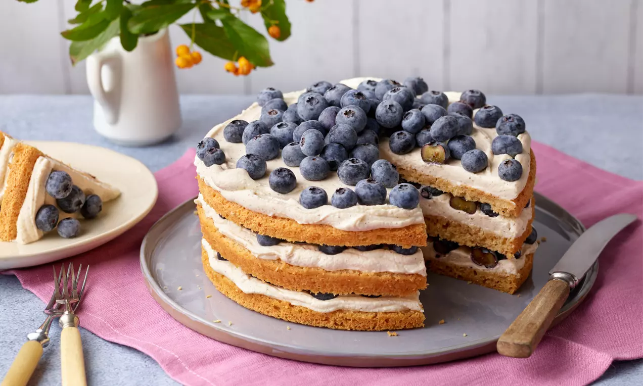 Mehrschichtige Torte mit hellbraunen Böden, heller Creme und frischen Heidelbeeren auf einem grauen Teller, daneben Messer und Gabel