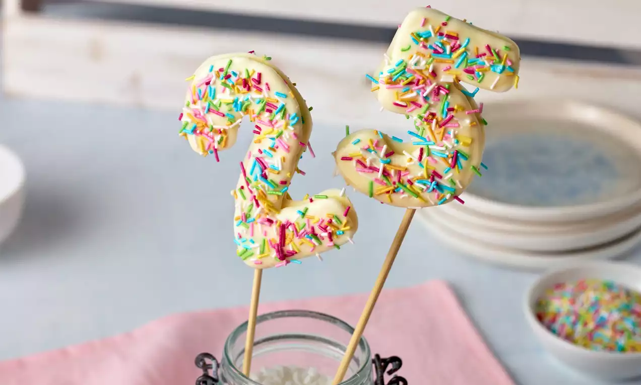 Zahl 25 aus weißer Schokolade auf Holzspießen, bedeckt mit bunten Streuseln, in einem Glas präsentiert auf rosa Stoff mit Teller im Hintergrund