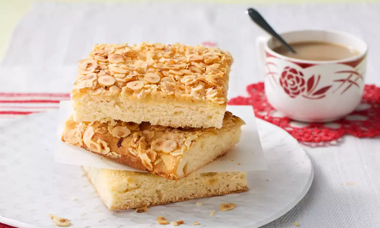 Butterkuchen mit goldgelber Kruste und gerösteten Mandelblättchen, rechteckig geschnitten und übereinander gestapelt auf einem weißen Teller