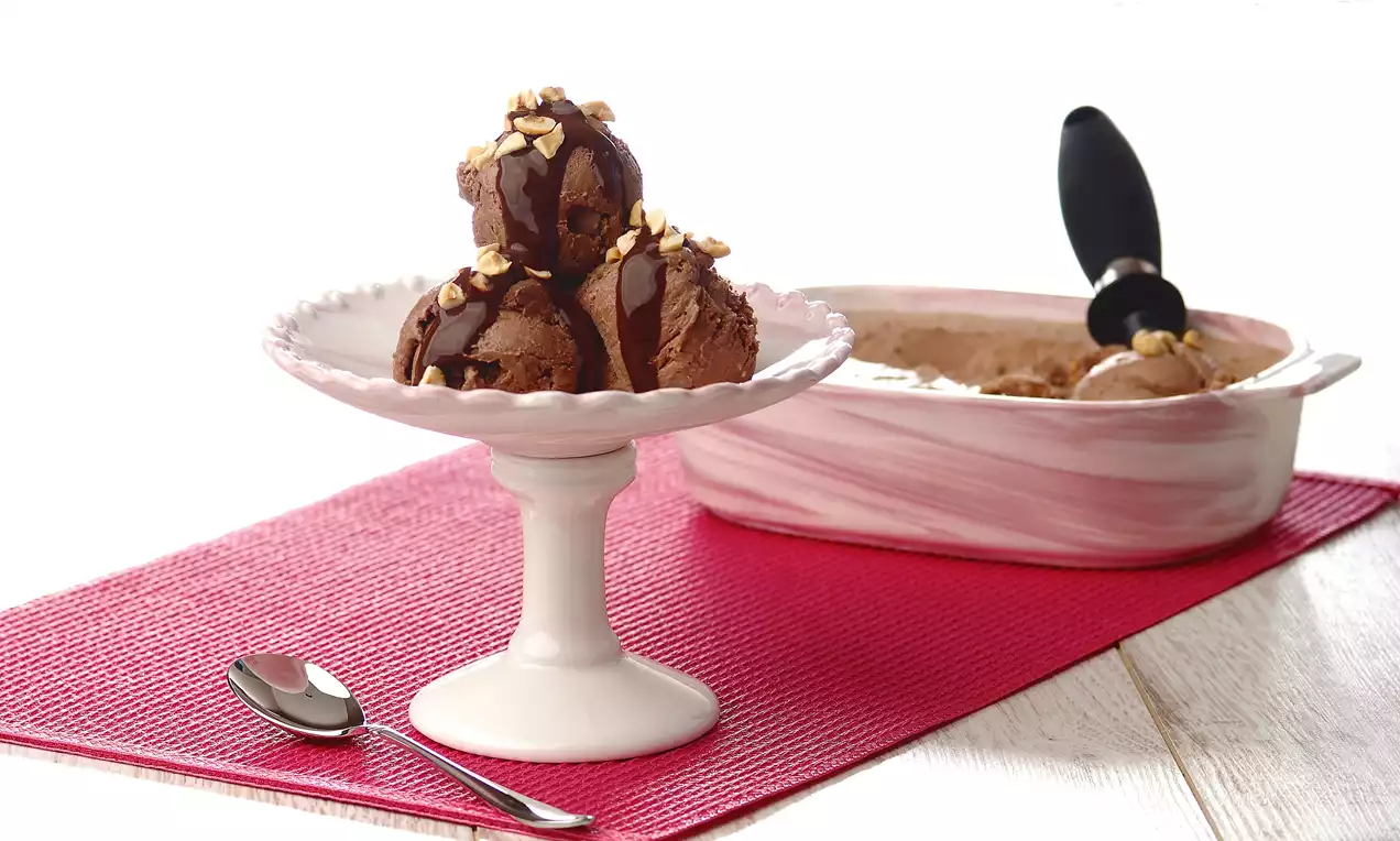 Kıvrımlı beyaz tabakta üç top çikolatalı dondurma, üzerine serpilmiş ince doğranmış fındıklarla, pembe doku üzerinde bir sunum
