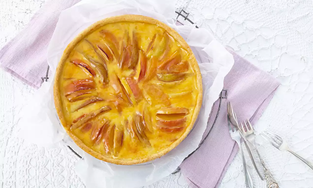 Französischer Obstkuchen mit goldgelber Glasur und dekorativ angeordneten Apfelscheiben auf weißem Teller mit lilafarbenen Servietten