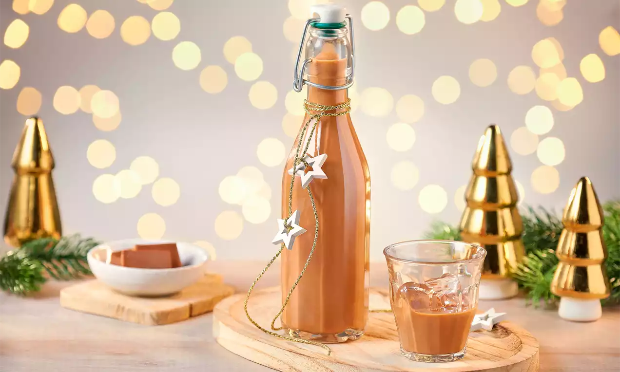 Karamel renkli likör şişesi yıldız süslerle dekore edilmiş, yanında cam bardakta likör ve altın detaylı yılbaşı dekorları bulunuyor