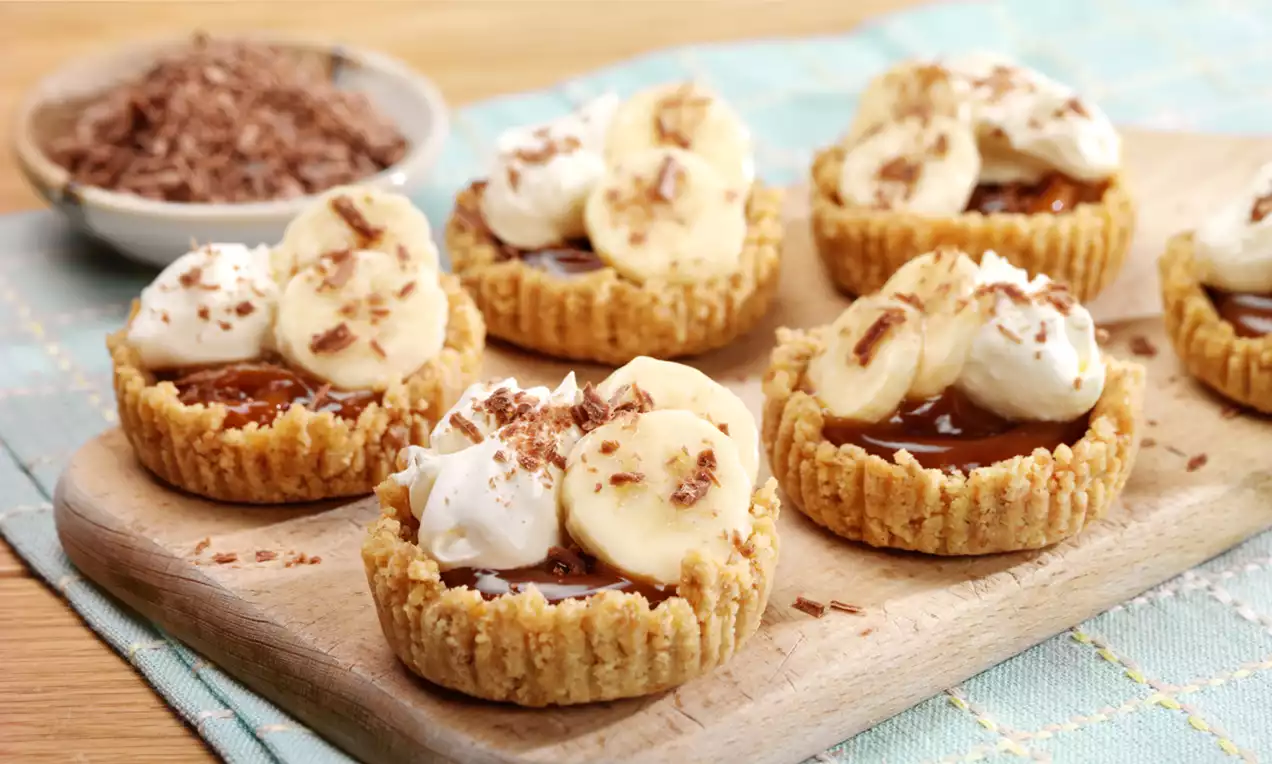 Mini banoffee pies with golden biscuit crusts, glossy caramel filling, banana slices, whipped cream, and chocolate shavings on a wooden board