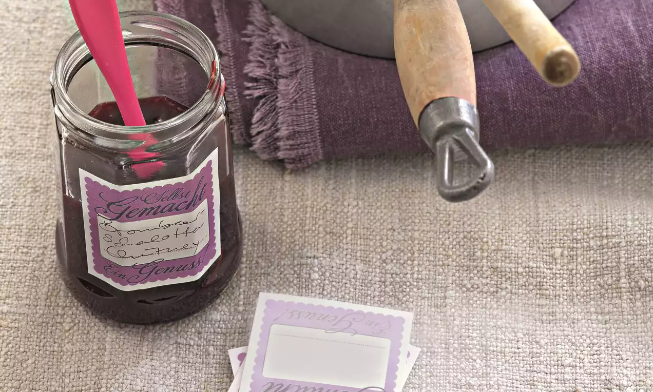 Ein Glas mit dunklem, samtigem Brombeer-Schalotten-Chutney, rosa Löffel, handbeschriftetes Etikett, auf beigem Tuch mit lila Stoff und Küchenutensilien