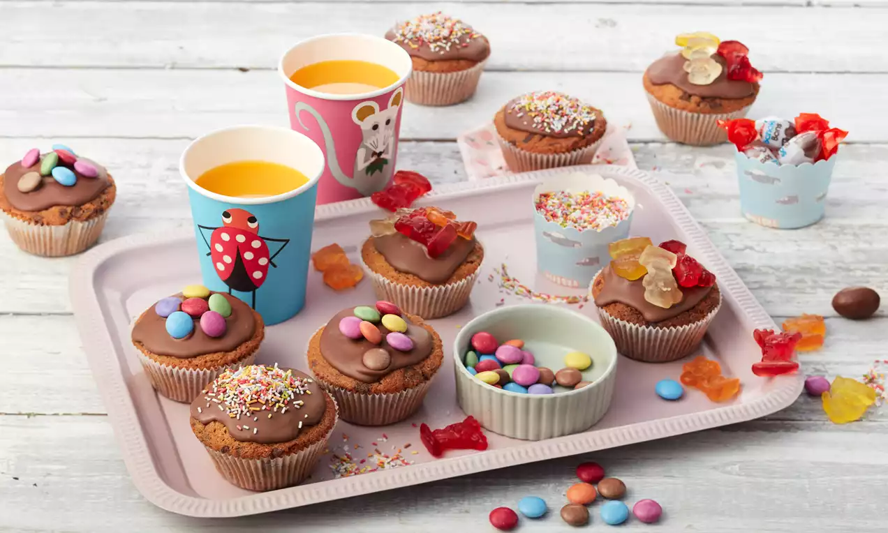 Schokoladenmuffins mit bunten Streuseln und Schokolinsen auf rosa Tablett, dekoriert mit Gummitieren, bunten Bechern und kleinen Schalen