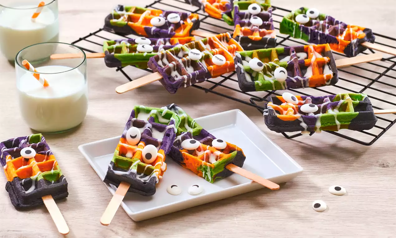 Waffe colorate în verde, portocaliu și mov, pe bețișoare, decorate cu ochișori din zahăr și glazură albă pe un grătar și farfurie albă