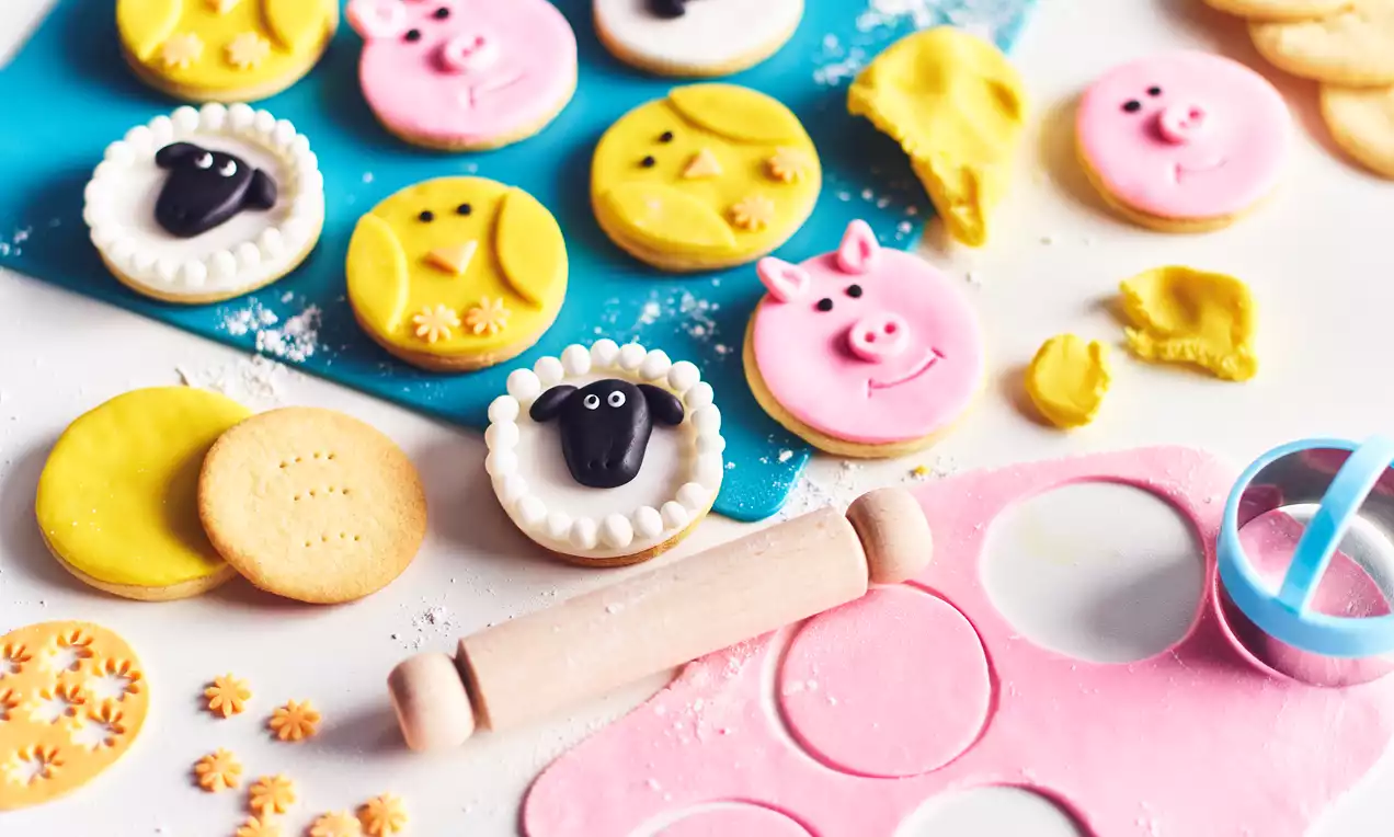 Animal-shaped cookies with pink, yellow, and white frosting, featuring pig, sheep, and smiley faces on a white and blue surface with baking tools