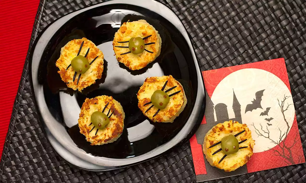 Petiscos dourados e gratinados decorados com azeitonas verdes e tiras pretas finas em um prato preto sobre jogo americano trançado escuro