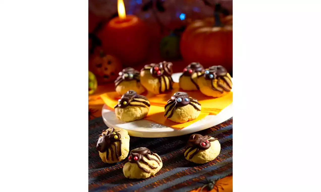 Spinnenkoekjes met beige ronde basis, chocoladestreams en kleurrijke snoepogen op een wit bord met oranje servet en Halloween-decoraties