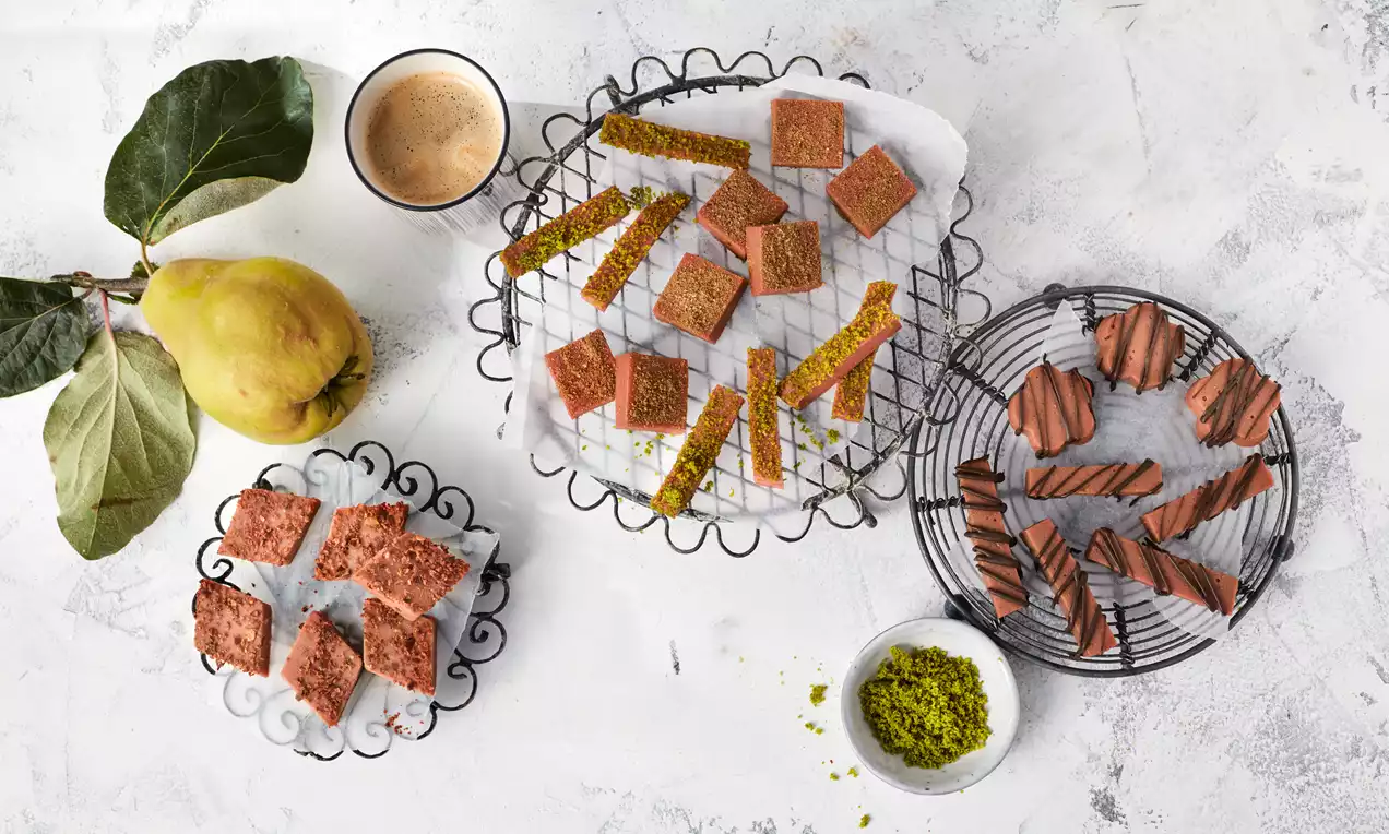 Quittenbrot in rotbraunen Quadraten und dünnen Streifen mit Pistazienrand auf Drahtgestellen, neben frischer Quitte und gemahlener Pistazie dekoriert