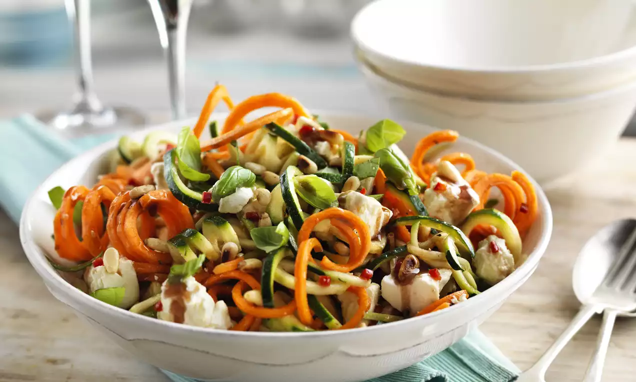 A white bowl filled with vibrant orange and green spiralised vegetables, feta cubes, fresh basil, and seeds, set on a pale blue napkin
