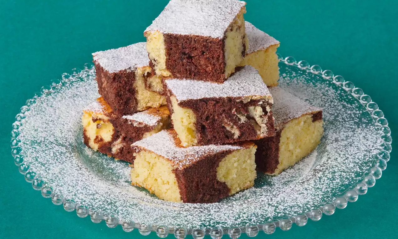 Kocky mramorového koláča s prepletenou žltohnedou textúrou, posypané práškovým cukrom, naaranžované na sklenenom tanieri.