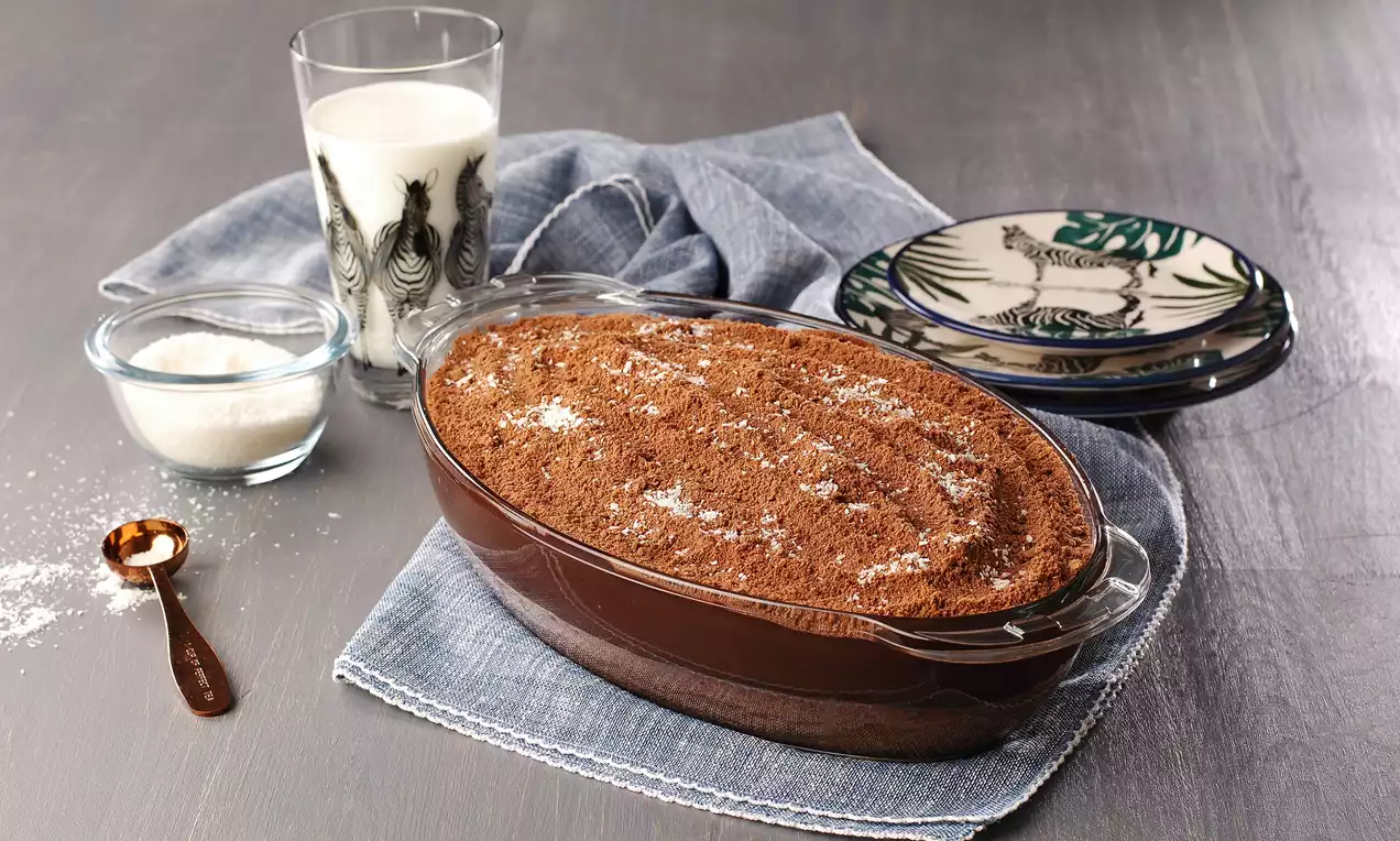 Koyu kahverengi oval cam kapta servise sunulan üzeri pudra şeker serpilmiş kumlu pasta, yanında bardakta süt ve dekorlu tabaklar eşlik ediyor