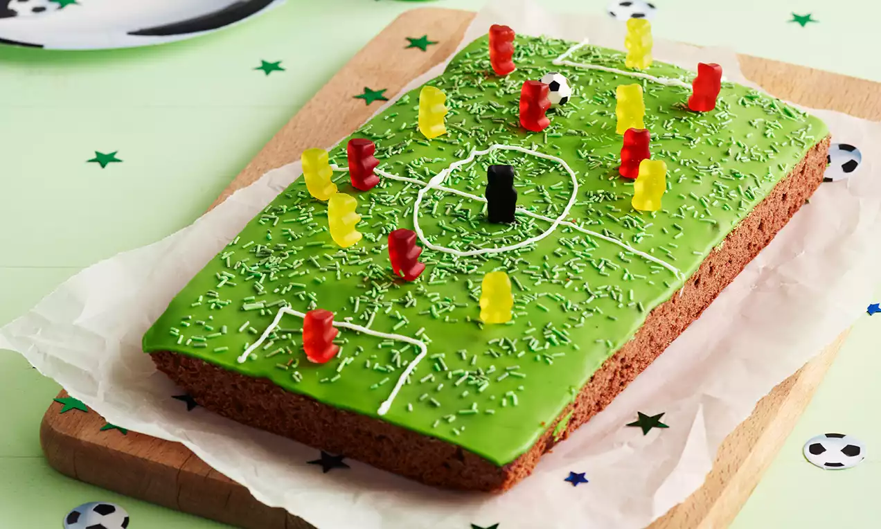 Rektangulær chokoladekage dækket med grøn glasur og hvide linjer som fodboldbane, pyntet med vingummifigurer og drys på bagepapir