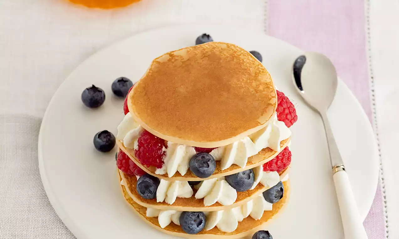 Gouden pannenkoekstapels met witte slagroom, blauwe bessen en frambozen op een wit bord met een zilveren lepel ernaast