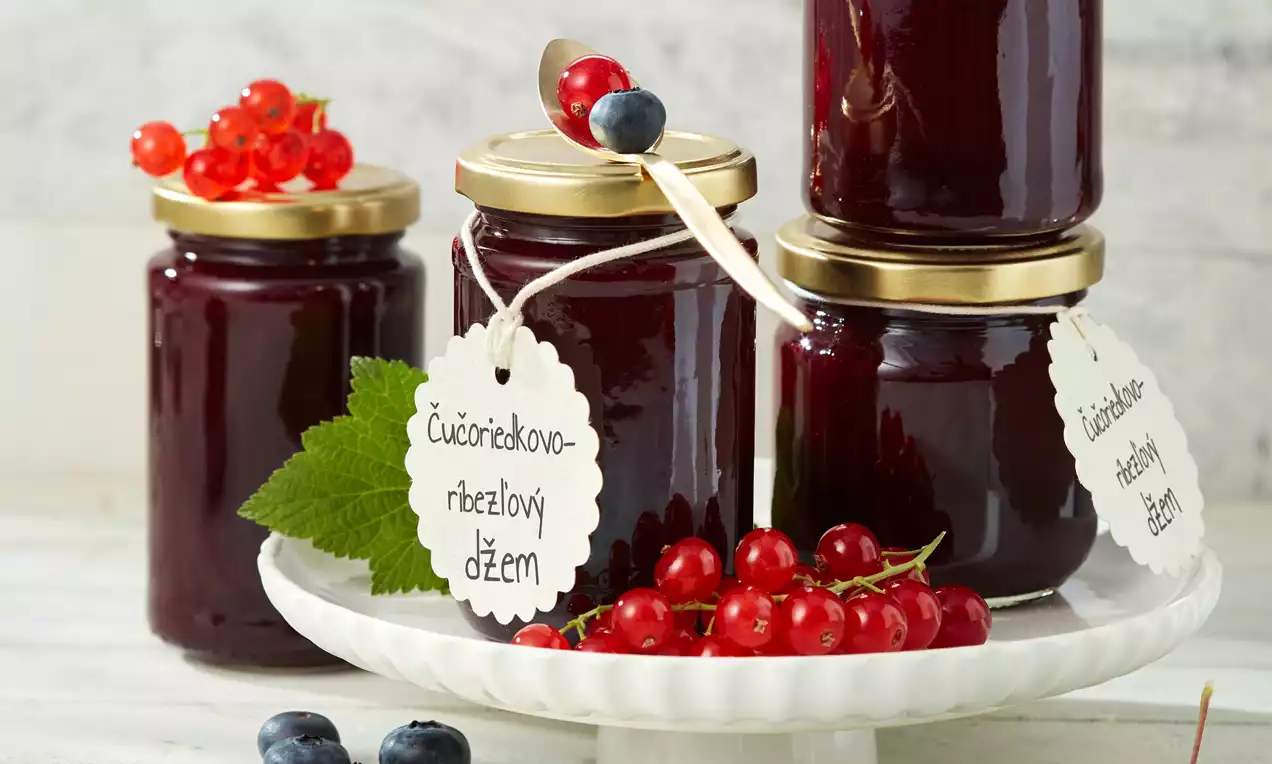 Čučoriedkovo-ríbezľový džem tmavorubínovej farby v sklenených pohároch so zlatými viečkami na bielom podnose, ozdobený čerstvými ríbezľami a čučoriedkami