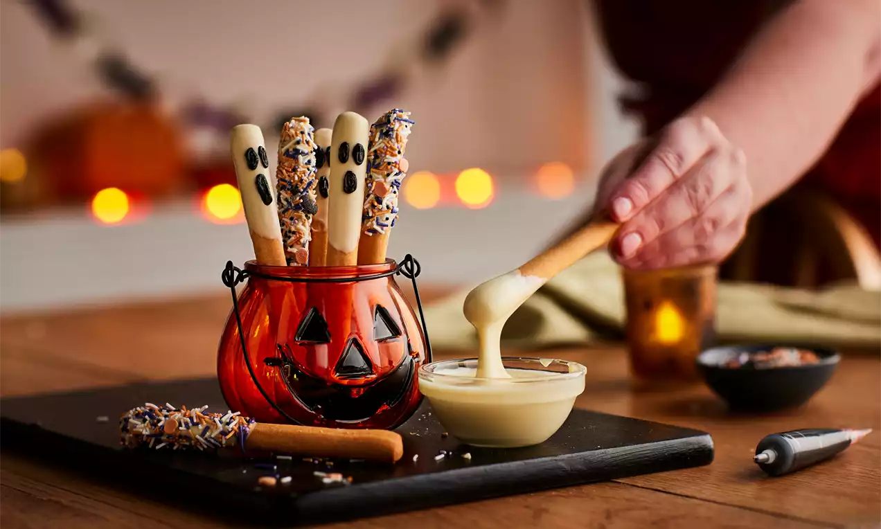 Ghost-shaped white chocolate-dipped breadsticks with black faces and sprinkled ends arranged in a red jack-o'-lantern pot on a table