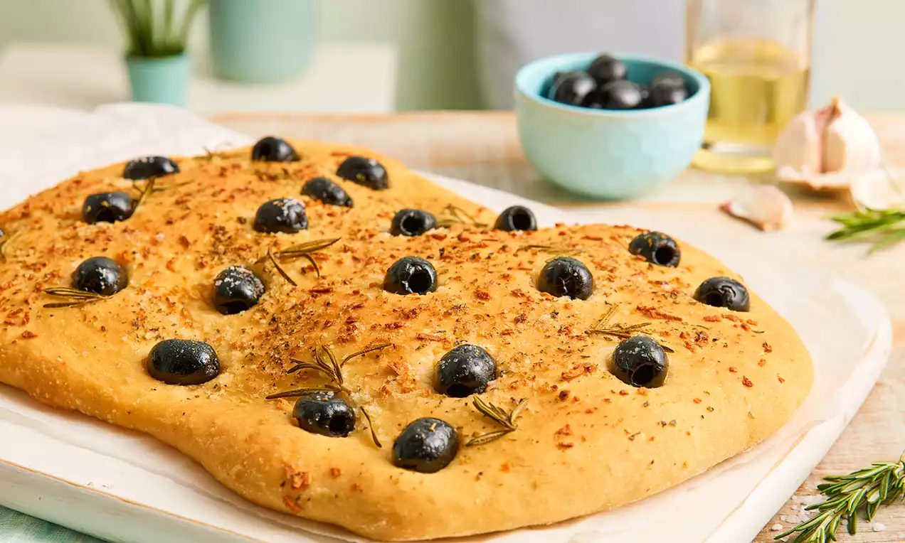 Goldene Focaccia mit schwarzem Olivenbelag, duftenden Rosmarinzweigen und fein verstreuten Kräutern auf einem weißen Teller mit Holzunterlage