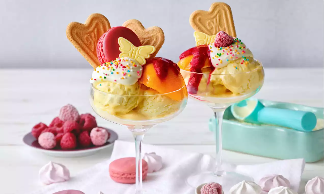 Pfirsich Melba in gläsernen Dessertschalen mit gelbem Eis, Pfirsichspalten, Himbeeren, Sahne, bunten Streuseln, Macaron und Gebäck verziert