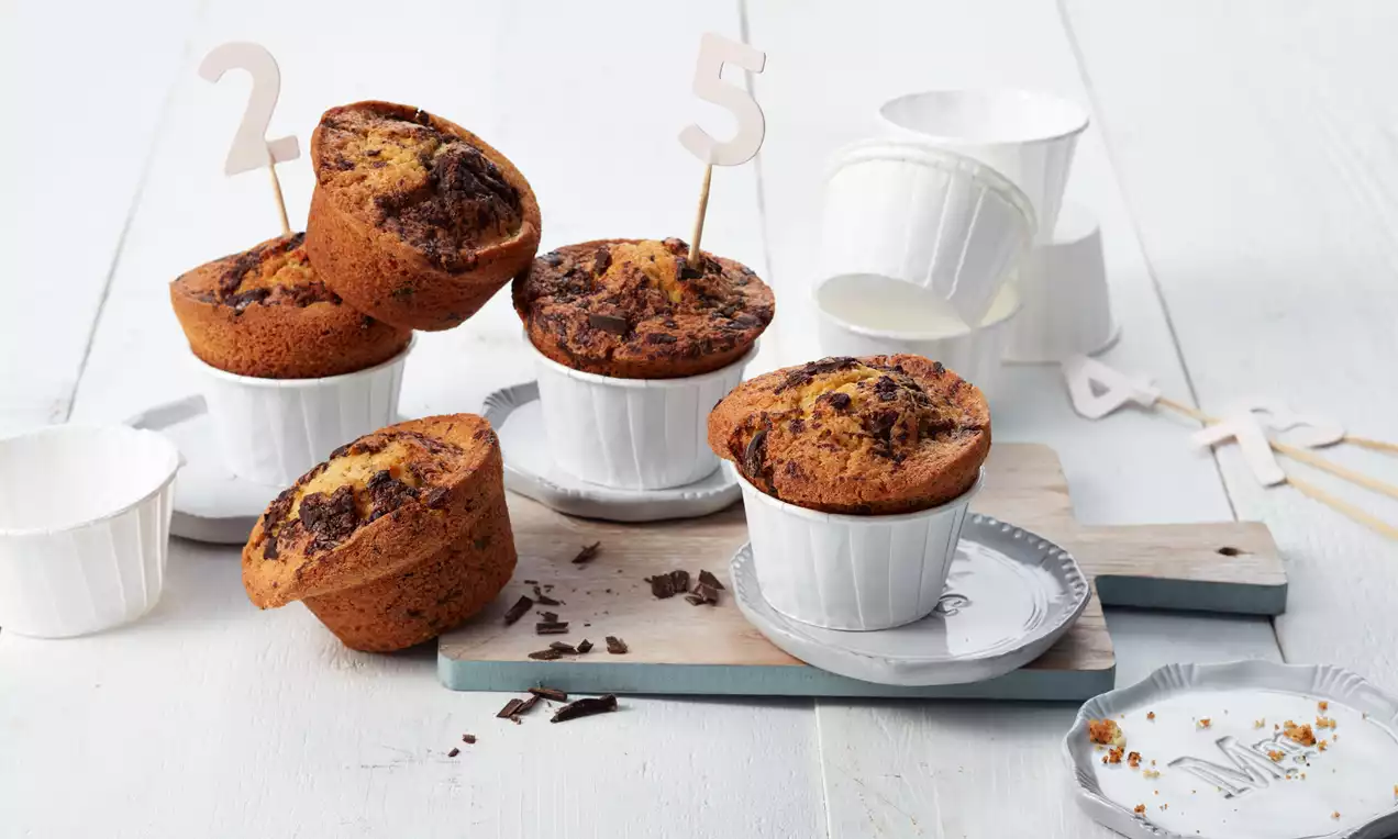 Goldbraune Muffins mit dunklen Schokosplittern in weißen Förmchen, arrangiert auf einem Holzbrett mit Zahlensteckern und Dekoration