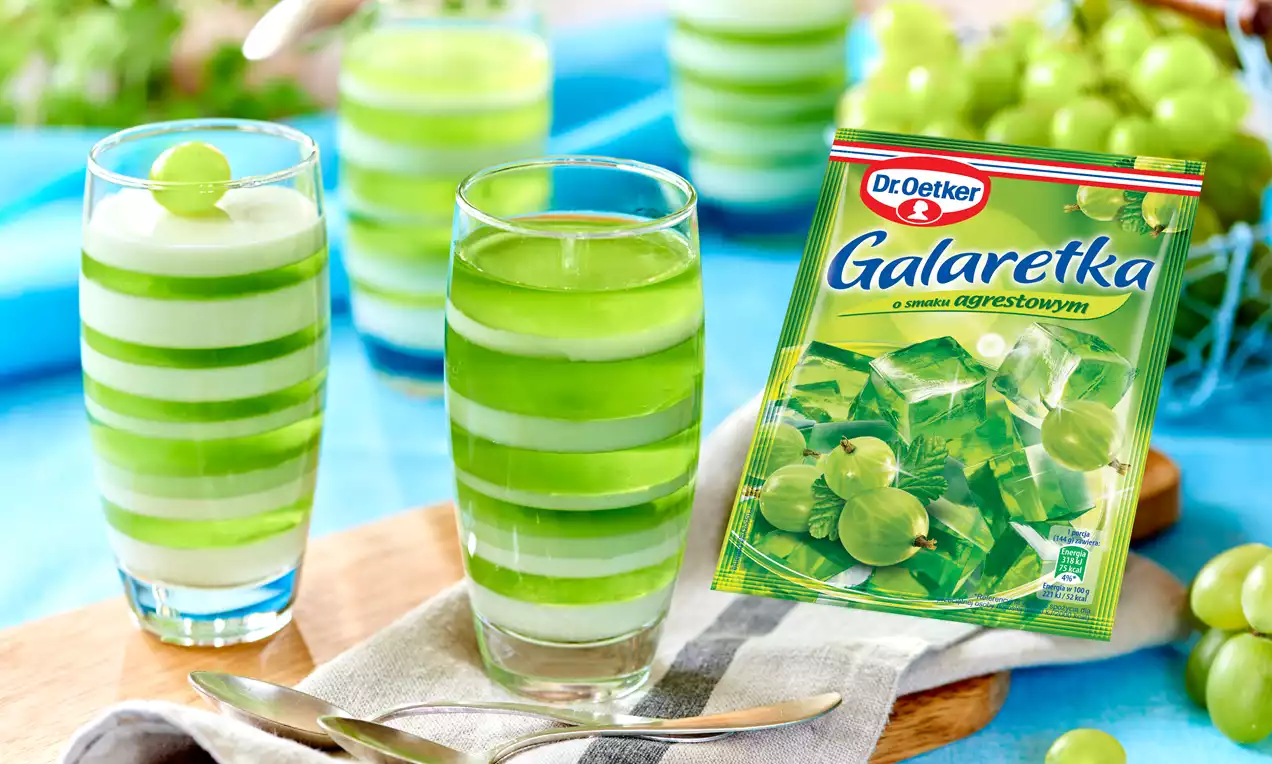 Warstwowy deser w wysokich szklankach z zieloną galaretką i kremem, udekorowany winogronami, obok łyżeczka na drewnianej desce i winogrona