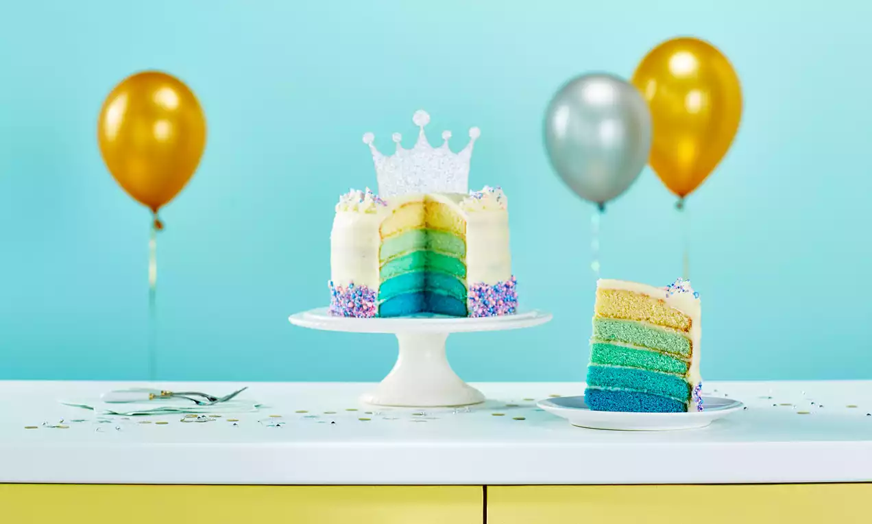 A layered cake with blue to yellow gradient sponge, white frosting, sprinkles, a glittery crown topper on a stand, and a slice on a plate