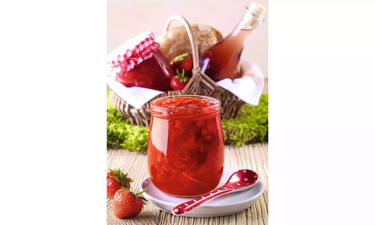 Sklenice zářivě červeného jahodového topping s lesklou strukturou, na talíři s puntíkovanou lžičkou, vedle čerstvých jahod a piknikového košíku